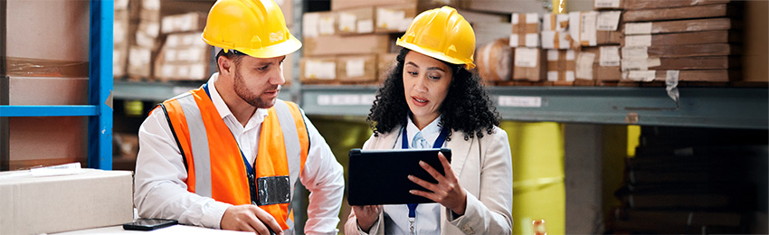 Two people wearing safety vests and hardhats in a warehouse reviewing a tablet.