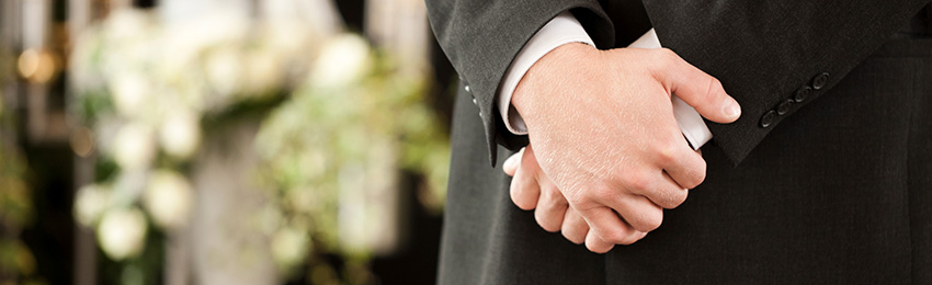 Politely clasped hands of a funeral director in front of some funeral bouquets.