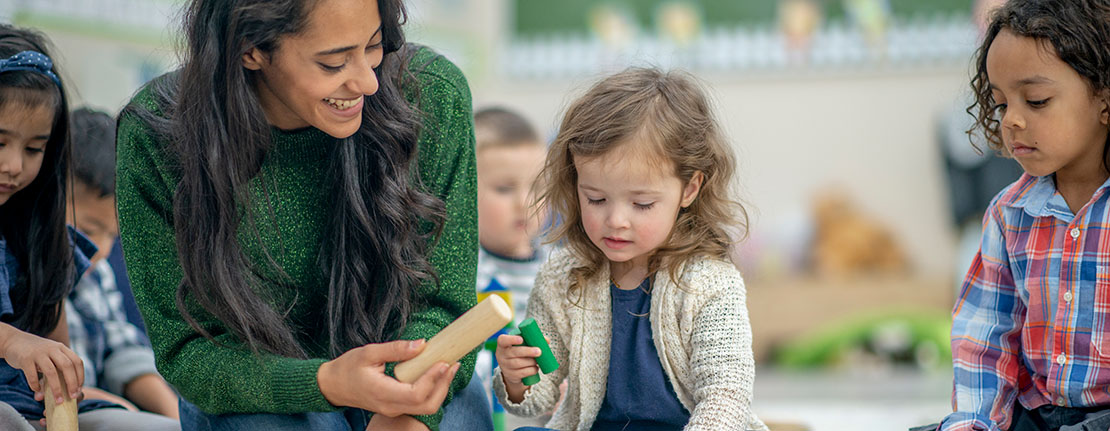 A teacher is seated next to preschool age children, helping them play with their blocks.
