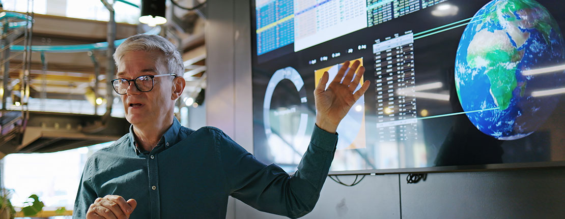 A man presenting in front of a screen displaying graphs, data and an image of the earth.
