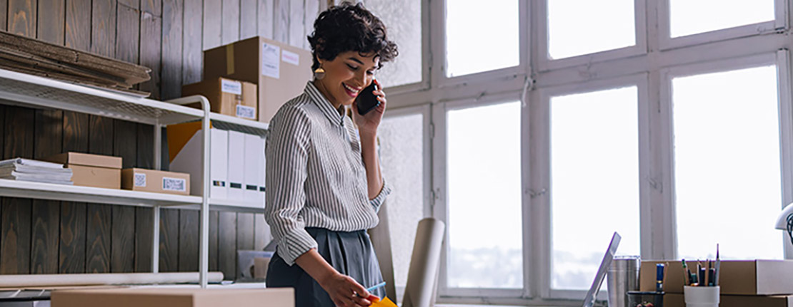 An energetic young woman in a professional office setting, happily engaged in a phone conversation. Surrounding her are a well-organized desk with a sleek laptop, indicating her entrepreneurial spirit, and a stack of shipping packages, showcasing her active business operations.